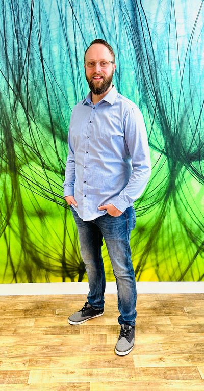 lächelnde Person mit beiden Händen in den Hosentaschen vor einer grünen Fototapete